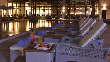 Zona de descanso junto a la piscina con tumbonas, una mesa con bebidas y aperitivos, y vistas a la piscina y a un área de estar interior.