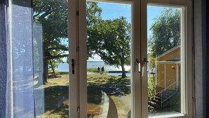 A window with a view of a lake, a wooden building, and a sailboat model. - Lövudden Strand (Vasteras)