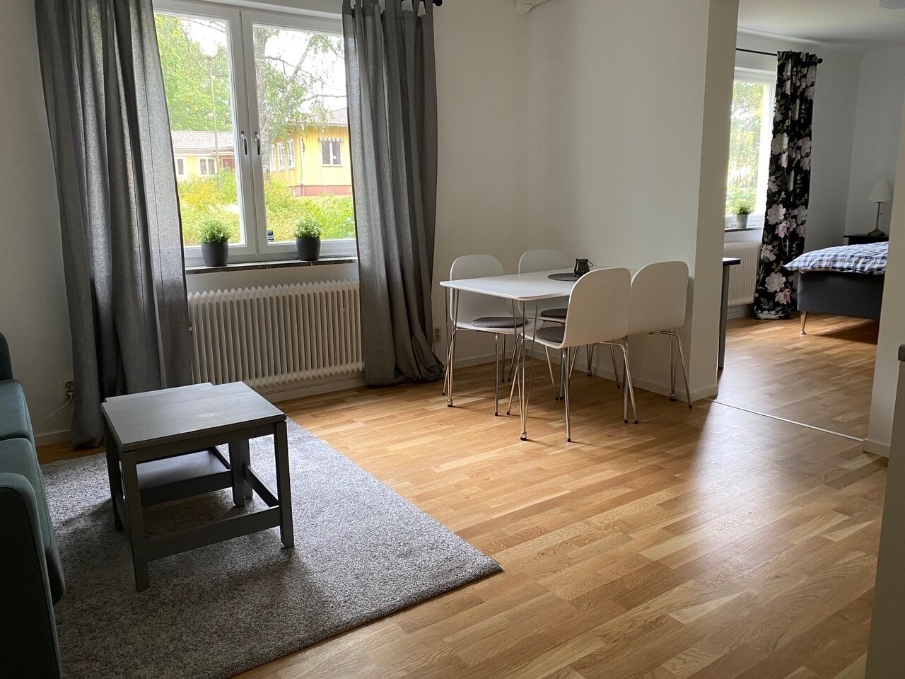 Ett rum med ett matbord och stolar, ett litet bord, ett element och ett fönster med gardiner.