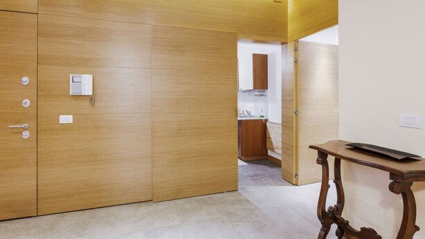 A room with a wooden wardrobe, a small table, and a kitchen area in the background. - Hostmate - Milano - Sapeto- Colonne S.Lorenzo (Milano)
