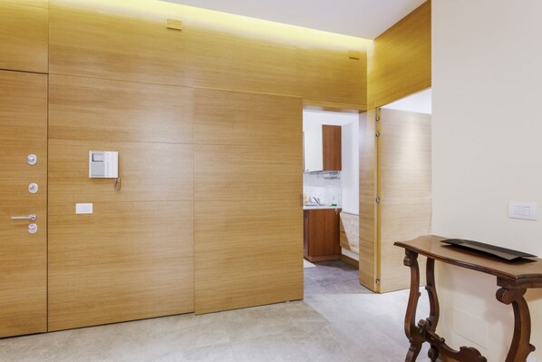 A room with a wooden wardrobe, a small table, and a kitchen area in the background.