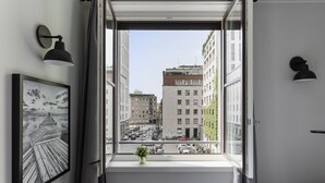 Een open raam met uitzicht op stadsgebouwen, een ingelijst kunstwerk aan de muur en een potplant op de vensterbank.