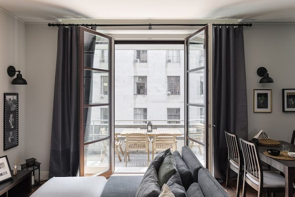 A room with a bed, a sofa, a dining table, and a view through a large window.