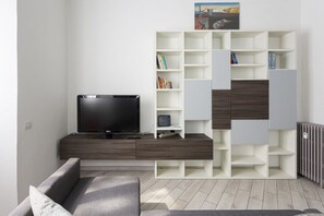 A modern living room with a flat-screen TV, a grey sofa, and a white shelving unit with books and decorative items. - Hostmate - Milano - Via Pacini- Lambrate (Milano)