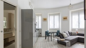 Uma sala de estar moderna com um sofá cinzento, uma mesa de jantar com cadeiras e uma área de cozinha com frigorífico.
