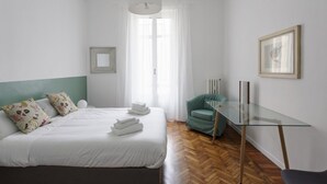 Habitación de hotel con una cama, una mesita de vidrio, una silla, una ventana con cortinas y un cuadro enmarcado en la pared.