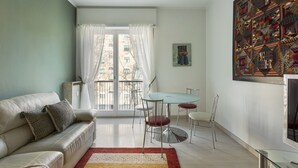 A living room with a sofa, a dining table with chairs, a large window with curtains, and a painting on the wall. - Hostmate - Milano - Cenisio - Bullona (Milano)