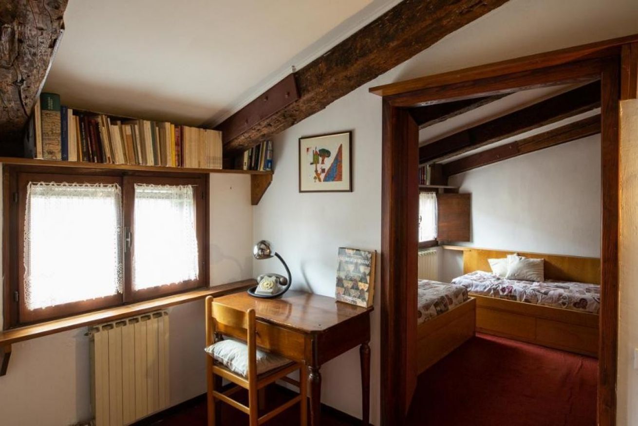 A room with a wooden desk, a chair, a bed, a radiator, and a window with lace curtains.