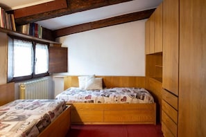 A small bedroom with two wooden beds, a window with lace curtains, and a bookshelf.