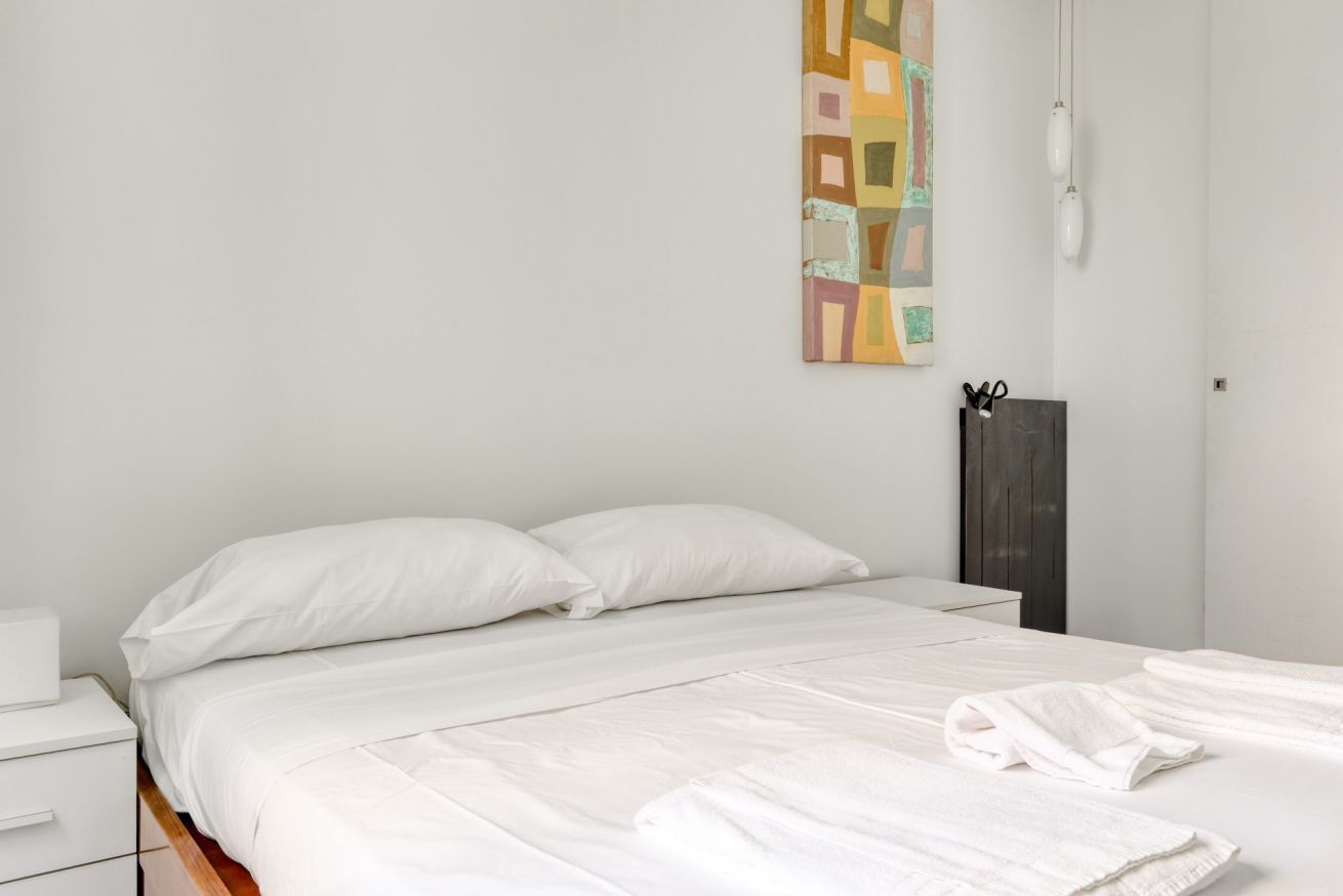 Un letto rifatto con lenzuola bianche, un comodino con una lampada e un'opera d'arte appesa al muro.