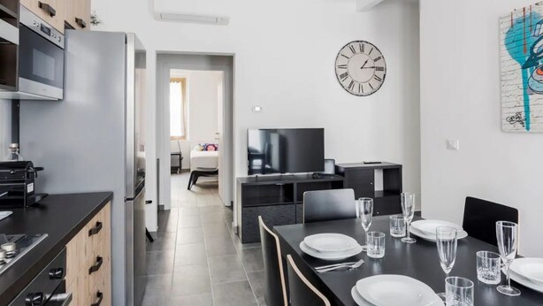 Eine moderne Küche mit Einbauofen und Mikrowelle, ein Essbereich mit einem schwarzen Tisch, gedeckt für vier Personen, und eine an der Wand befestigte Uhr.