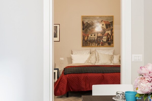 A bedroom with a bed, a painting on the wall, a table with a vase, and a chair.