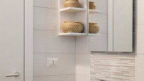 Un bagno con pareti rivestite di piastrelle bianche, una porta bianca, uno specchio e mensole con cesti in vimini.