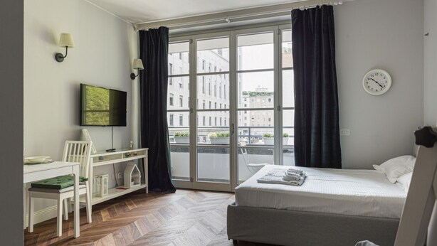 A room with a bed, a desk with a chair, a television, and a window with curtains. - Hostmate - Milano - Freguglia's Studio - Guastalla (Milano)