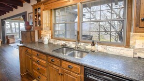 A kitchen with wooden cabinets, a granite countertop, a double sink, and a large window with a view of trees. - Grandfather Mountain Views, Spacious Deck & Game Room Near Blowing Rock (Blowing Rock)