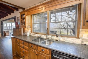 A kitchen with wooden cabinets, a granite countertop, a double sink, and a large window with a view of trees. - Grandfather Mountain Views, Spacious Deck & Game Room Near Blowing Rock (Blowing Rock)