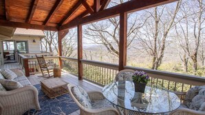 Une terrasse extérieure couverte, aménagée avec des meubles en osier, une table en verre et offrant une vue sur les arbres et les collines.