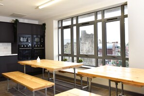 A modern kitchen with built-in appliances, large windows with a view of a historic building, and a wooden table with benches. - Cwtsh Hostel (Swansea)