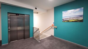 Un ascenseur doté d’un écran numérique, un escalier muni d’une rampe et une peinture représentant un paysage urbain accrochée au mur.