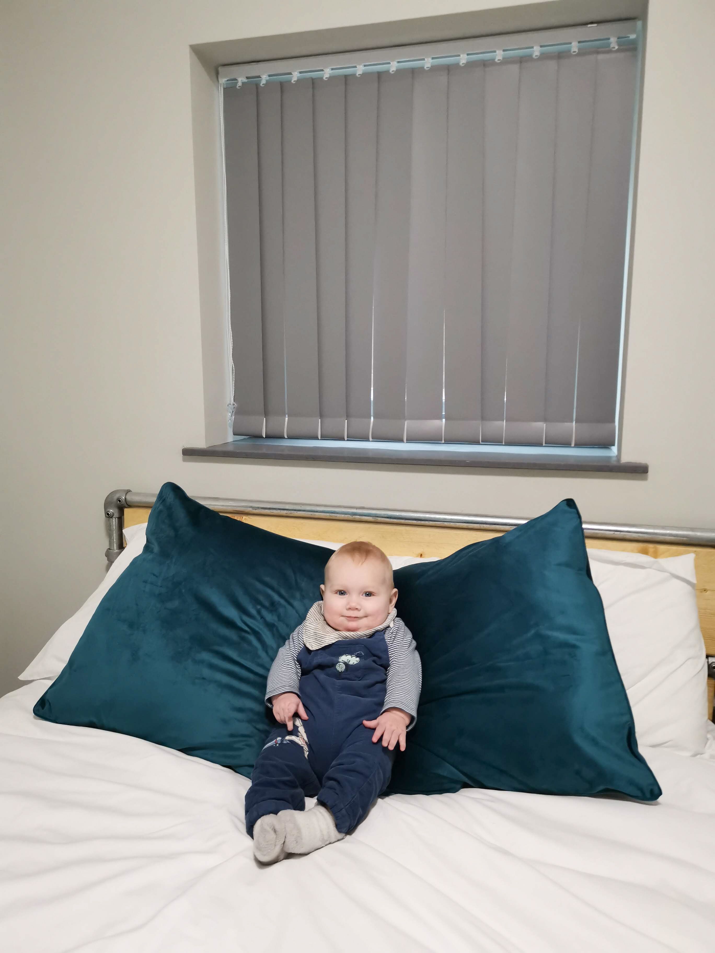 A child sitting on a bed with two large pillows, a window with blinds above, and a metal headboard.