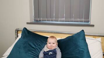 A child sitting on a bed with two large pillows, a window with blinds above, and a metal headboard.