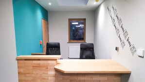 Un comptoir d’accueil avec deux chaises noires, un bureau d’accueil en bois, une fenêtre avec des stores et un mur orné d’un motif textuel répété.