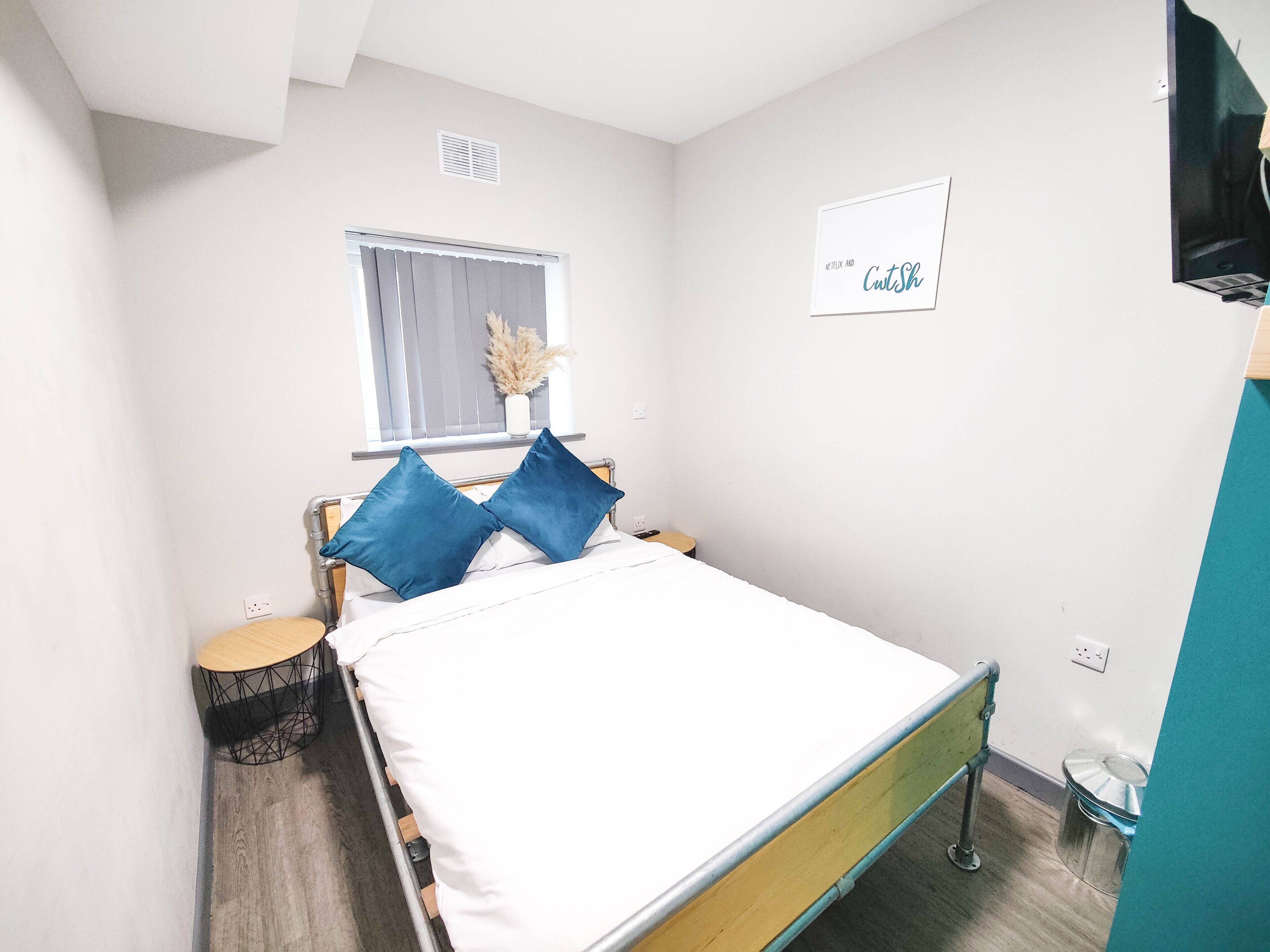 Een slaapkamer met een bed, een nachtkastje, een raam met zonwering, een tv aan de muur en een bordje met de tekst 'Netflix and CutSh'.