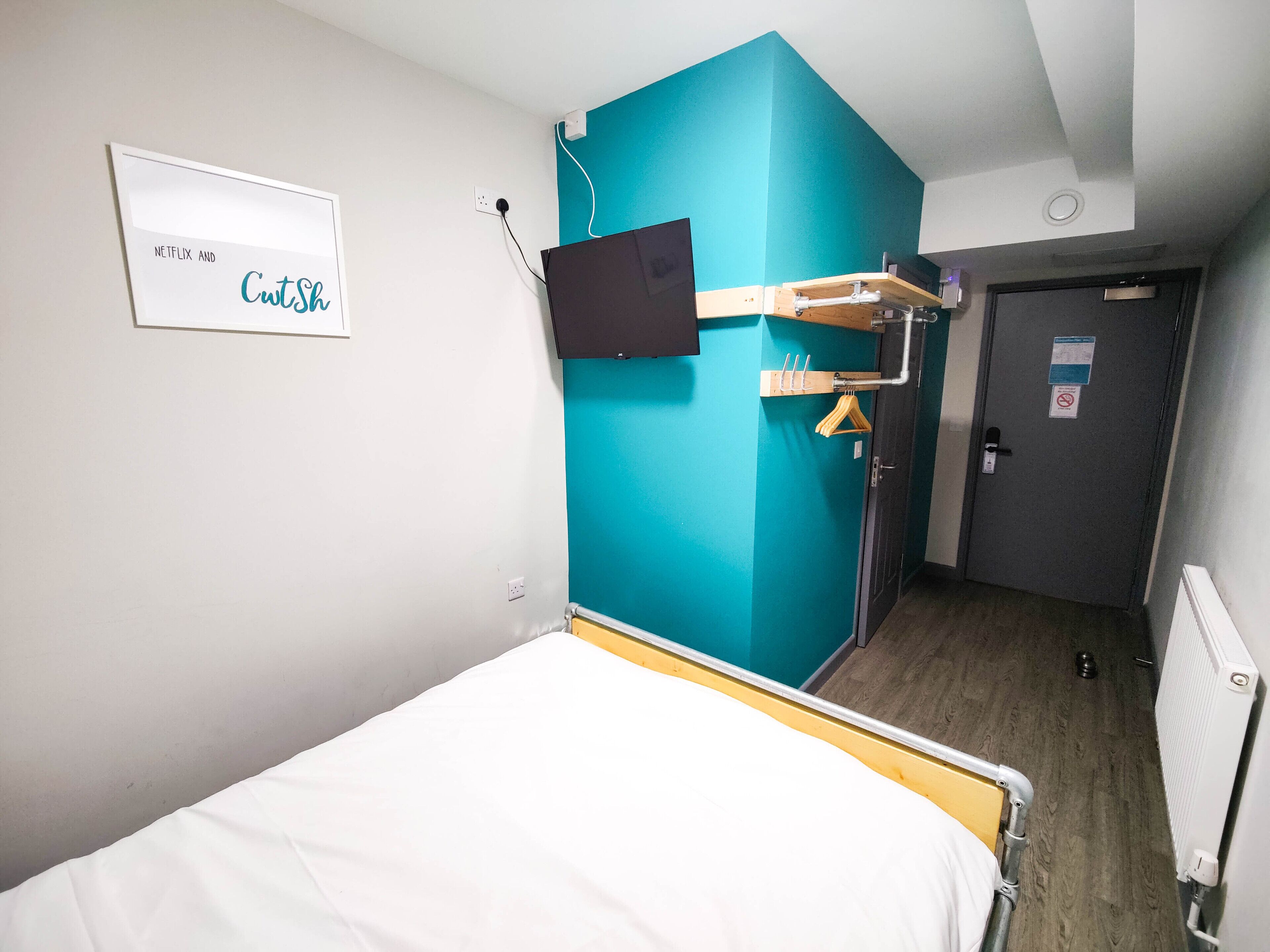 A hotel room with a bed, a TV mounted on the wall, a teal accent wall, and a door with a sign.