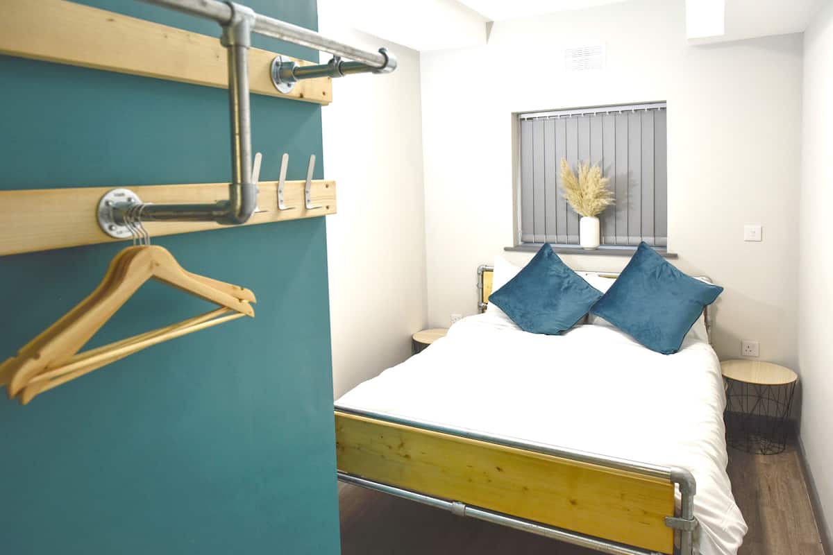 A bedroom with a bed, a wooden hanger, a window with a plant, and a small round table.
