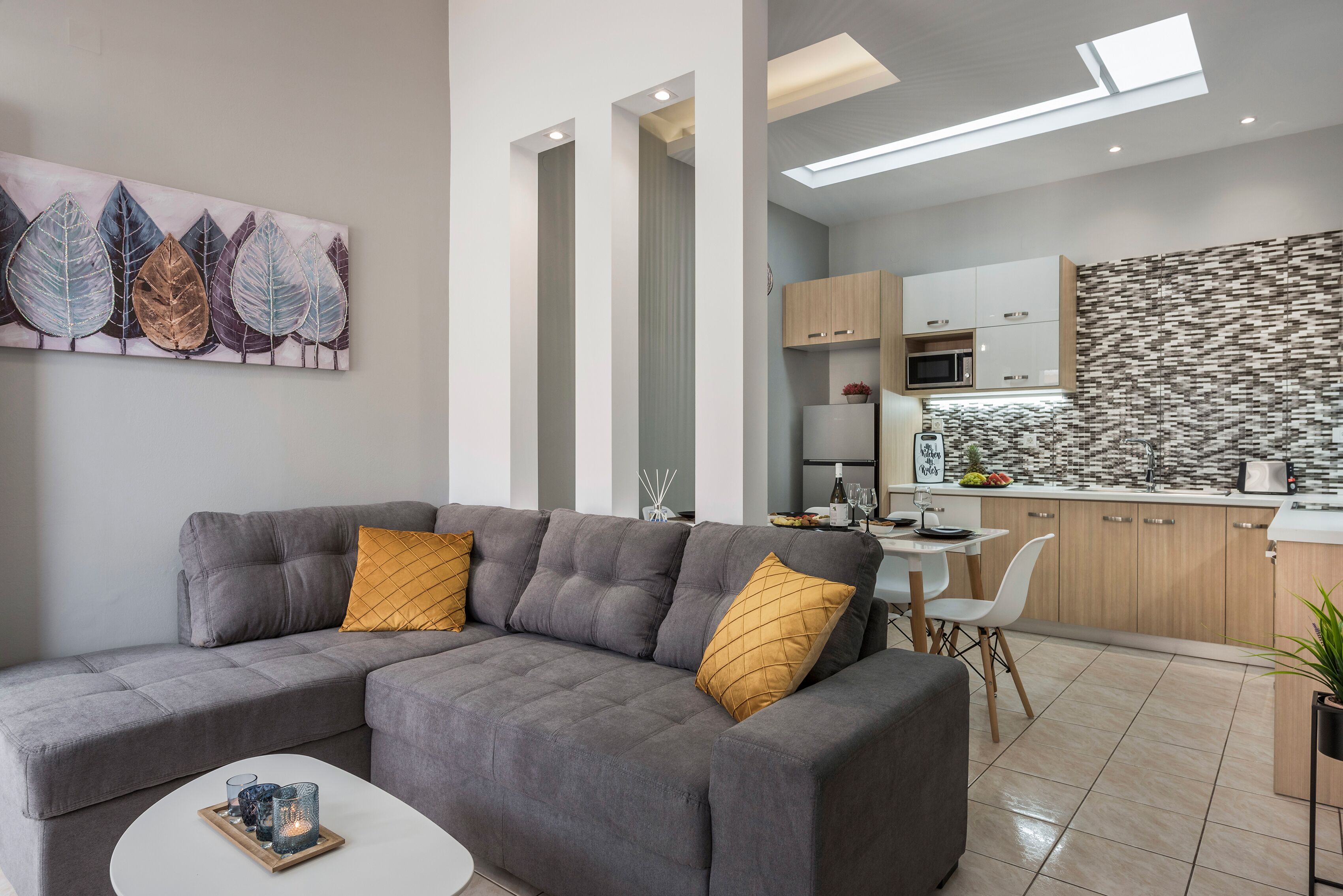 Ein modernes Wohnzimmer mit einem grauen Ecksofa, einem kleinen weißen Couchtisch und einem Blätterbild an der Wand.