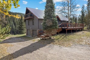 A two-story cabin with a balcony, surrounded by trees and a forested hillside.