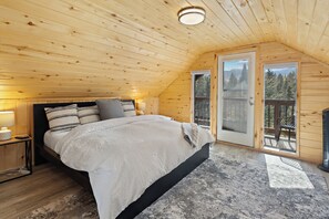A bedroom with a wooden ceiling, a bed with a white comforter, a nightstand with a lamp, and a view of a balcony with a wooden railing.