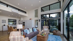 A spacious living room with a large sofa, a coffee table, and a chair. There is a kitchen area with a dining table and chairs visible in the background. - KAURI HILL ESTATE Luxe Retreat Overlooking Whangaroa Harbour (Northland)