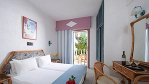 A hotel room with a bed, a desk with a chair, a mirror, and a balcony with a view of greenery. - Fan Village (Hersonissos)