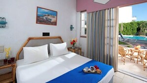 A hotel room with a bed, a small table with a vase of flowers, a painting on the wall, and a view of a pool and outdoor seating area. - Fan Village (Hersonissos)