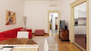 A living room with a red sofa, a wooden coffee table, a TV on a wooden stand, and a kitchen visible through an open door. - Hostmate - Milano - Ozieri (Milano)