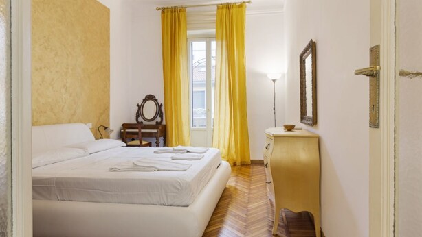 A bedroom with a bed, a dresser, a mirror, and a window with curtains. - Hostmate - Milano - Ozieri (Milano)