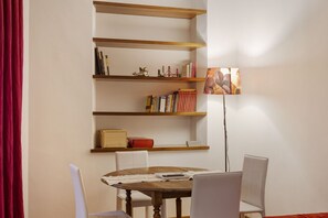 Una habitación con una mesa de madera, sillas blancas, una estantería con libros y objetos decorativos, y una lámpara de pie.