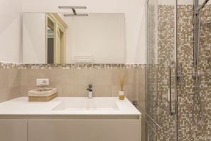 A bathroom with a white sink, a mirror, a shower with mosaic tiles, and a wicker basket on the counter.