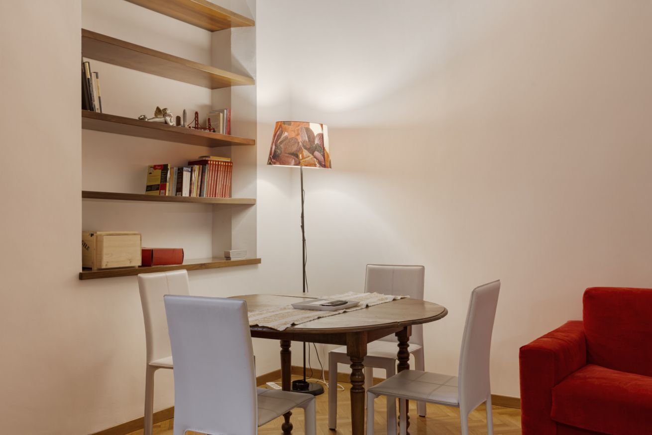 A dining area with a round table, white chairs, a red sofa, and wooden shelves with books and decorative items.
