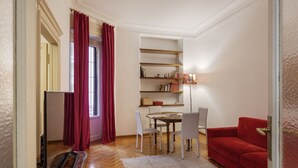 A living room with a red sofa, a dining table with chairs, a bookshelf, and a window with red curtains. - Hostmate - Milano - Ozieri (Milano)