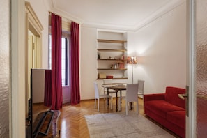 A living room with a red sofa, a dining table with chairs, a bookshelf, and a window with red curtains.