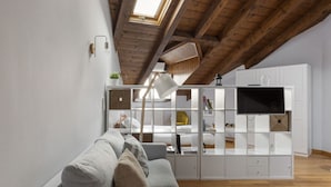A modern loft-style room with a high ceiling, exposed wooden beams, a built-in white entertainment unit, and a grey sofa. - Hostmate - Milano - Farini - Garibaldi (Milano)