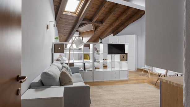 A modern loft-style room with a high ceiling, exposed wooden beams, a built-in white entertainment unit, a gray sofa, and a large window. - Hostmate - Milano - Farini - Garibaldi (Milano)