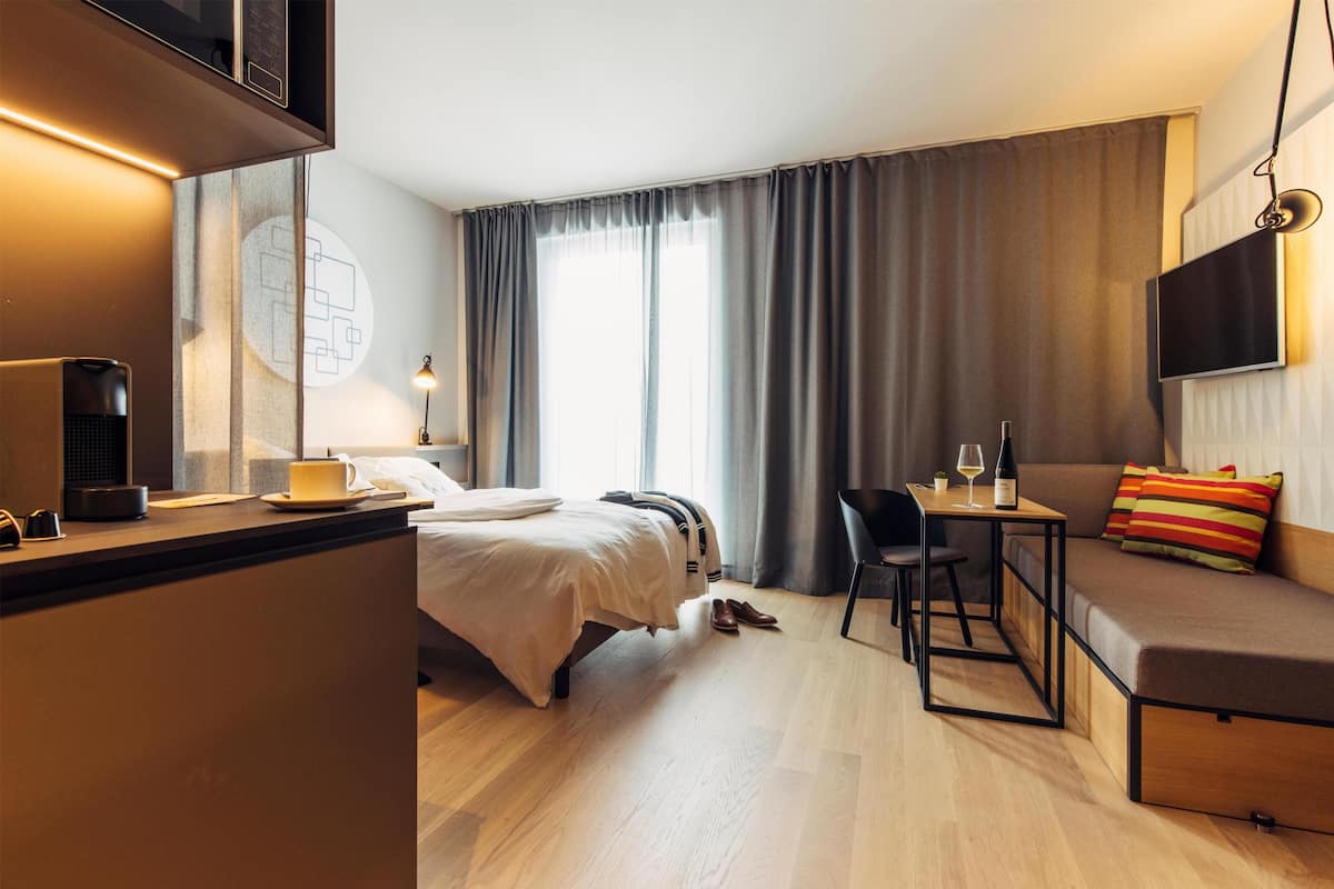 Ein modernes Hotelzimmer mit einem Bett, einem Schreibtisch, einer Couch, einem Fernseher und einem großen Fenster mit Vorhängen.