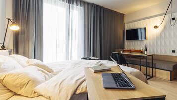 Quarto de hotel moderno com cama, escrivaninha com laptop, televisão e uma janela grande com cortinas.