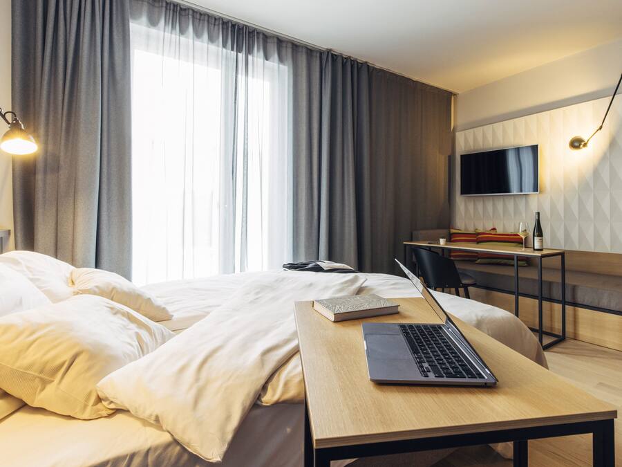 A modern hotel room with a bed, a desk with a laptop, a TV, and a large window with curtains.