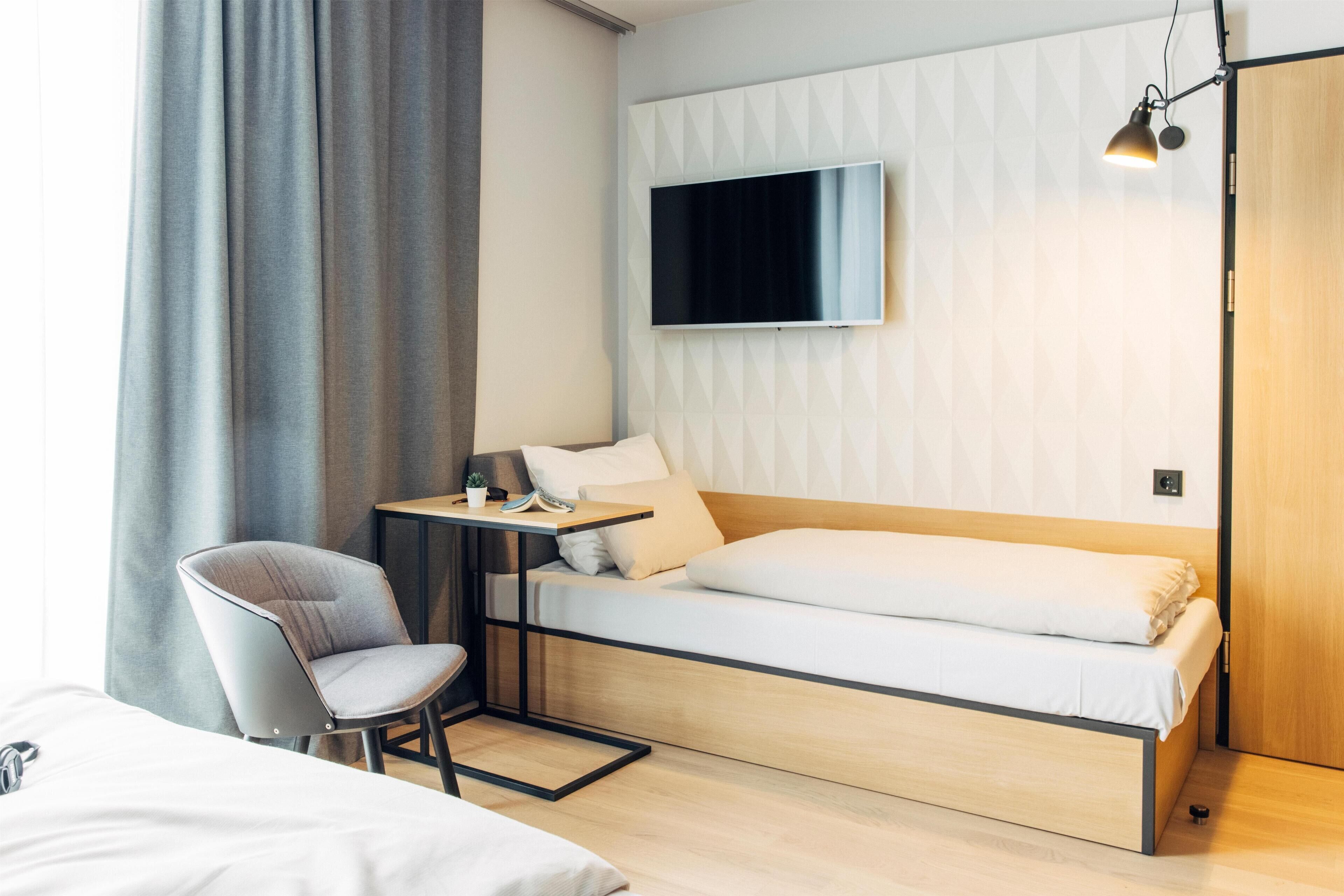 A modern hotel room with a bed, a desk with a chair, a TV mounted on the wall, and a wooden door.