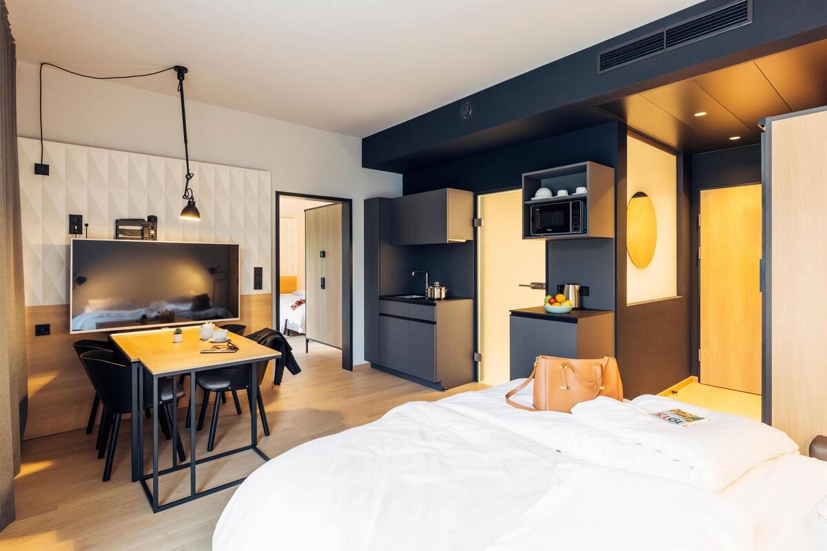 Ein modernes Hotelzimmer mit Bett, Schreibtisch mit Stühlen, einer Küchenzeile und einem Fernseher.