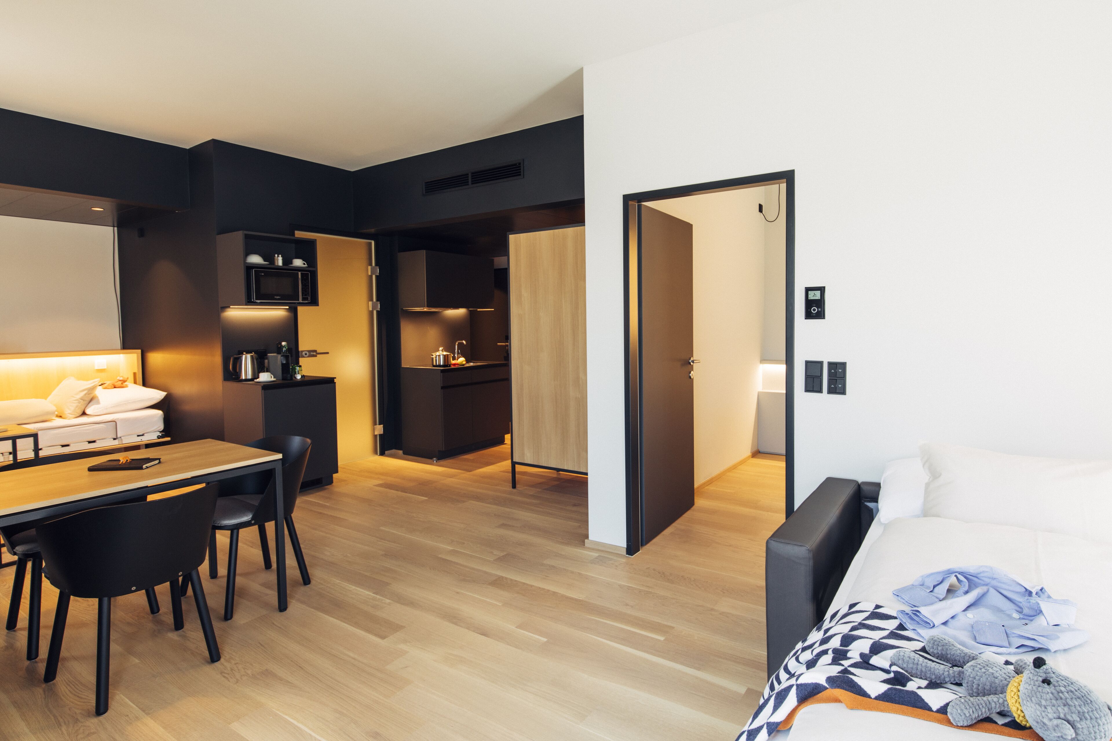 A modern hotel room with a wooden floor, a dining area with a table and chairs, a kitchenette, and a bed with a blanket and stuffed toy.